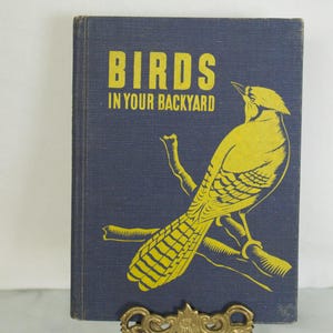 Może przedstawiać: Zabytkowa książka zatytułowana "Birds in Your Backyard" z żółtą ilustracją ptaka siedzącego na gałęzi na ciemnoniebieskim tle. Książka jest prezentowana na ozdobnym, złotym stojaku.