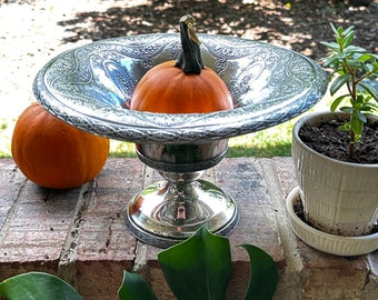 May include: A silver decorative bowl with an ornate design sits on a brick surface. A small orange pumpkin sits inside the bowl. A green plant in a white pot is to the right of the bowl.