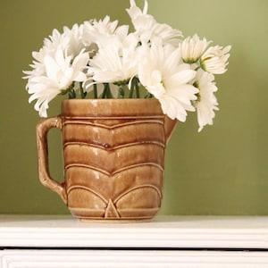 May include: A brown ceramic pitcher with a decorative design, filled with white daisies.