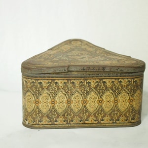 Puede incluir: Una caja de hojalata vintage triangular con un diseño estampado en dorado y marrón. La tapa está cerrada y la caja está sobre una superficie blanca.