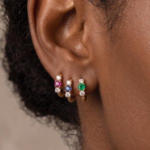 May include: Three small gold hoop earrings worn in a row on an earlobe. Each hoop features a central round gemstone in a different colour—pink, blue, and green—flanked by two small clear stones, creating a vibrant and colourful jewellery set.