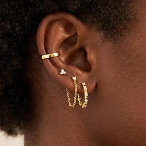 Può includere: Un ear cuff dorato, un piccolo orecchino a lobo con tre minuscole pietre chiare e un orecchino a cerchio dorato con una catenina sottile che si collega a un secondo cerchio, tutti con finitura in oro lucido e pietre chiare incastonate.
