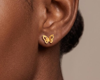Puede incluir: Un pequeño pendiente de botón dorado con forma de mariposa y alas texturizadas. El pendiente está colocado en el lóbulo de la oreja, presenta un acabado amarillo metálico y detalles intrincados en la superficie de las alas.