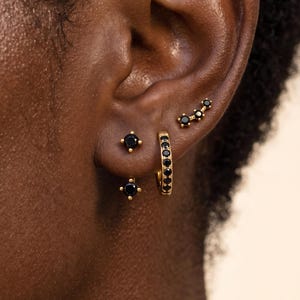 May include: A set of gold-toned earrings featuring black stones. The collection includes a small hoop earring with embedded black stones, a single stud earring with a black stone, and a curved ear climber earring adorned with four black stones.