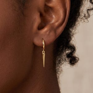 Op de afbeelding: Een gouden oorring met een kleine, bungelende spijkerbedel. De oorbel bestaat uit een dunne, cirkelvormige gouden ring die door de oorlel gaat, met een slanke, puntige gouden spijkerhanger die aan de onderkant van de ring is bevestigd.