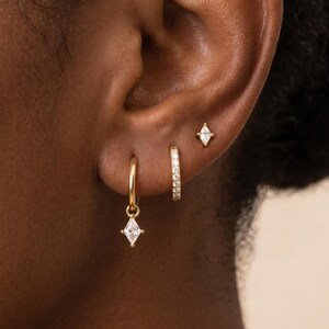 May include: A set of three gold-toned earrings consisting of a small hoop with a dangling marquise-cut clear stone, a thin pavé-set hoop, and a single marquise-cut stud earring. Each piece features a polished gold finish and clear stones.