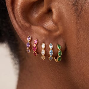 May include: Five small gold hoop earrings, each featuring three marquise-cut gemstones in a vertical row. The earrings display different colored stones, including purple, pink, clear, light blue, and green, set in a textured gold metal frame.