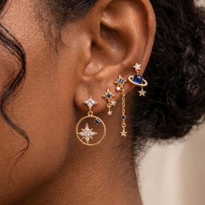 May include: A set of four gold-toned earrings featuring celestial motifs. The designs include a star inside a circular frame, a small star with a dangling chain, a star with a blue stone, and a planet with a blue sphere and a ring.