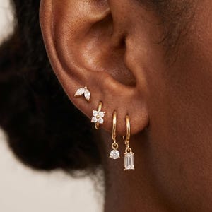 Könnte beinhalten: Ein Set aus vier goldenen Ohrringen an einem Ohr. Der oberste Ohrring ist ein Stecker mit marquise-geschliffenem Stein, gefolgt von einem kleinen Creolen-Ohrring mit Blumenanhänger, einem Creolen-Ohrring mit rundem Anhänger und einem mit Baguette-Stein.