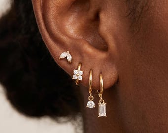 May include: A set of four gold earrings worn in a single ear. The top earring is a marquise-cut stone stud, followed by a small hoop with a four-stone flower charm, a hoop with a round solitaire drop, and a hoop with a rectangular baguette drop.