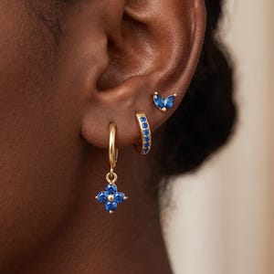 Peut inclure: Un ensemble de trois boucles d'oreilles dorées ornées de pierres bleues. L'une est un anneau avec une breloque fleur à quatre pétales, une autre est un petit anneau serti de pierres bleues, et la troisième est un petit clou en forme de feuilles.