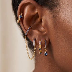 May include: A set of gold-toned earrings and ear cuffs featuring deep blue stones. The collection includes a beaded ear cuff with a round stone, a chain-linked ear cuff, and three small hoop earrings with blue stone accents and a teardrop charm.