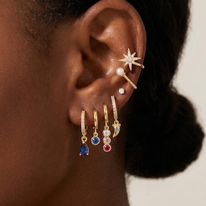 Può includere: Una collezione di orecchini dorati indossati su un orecchio, tra cui un ear cuff a forma di stella con pietre chiare, un ear cuff di perla, un piccolo perno e quattro orecchini a cerchio con una goccia blu, una pietra blu, un ciondolo multicolore e un corno.