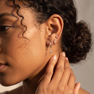 May include: A set of three silver-toned earrings worn on an earlobe. The jewelry includes one small stud earring with a star-shaped design, one plain silver hoop, and one silver hoop featuring a dangling star-shaped charm with a central point.