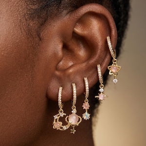 May include: A set of five gold hoop earrings featuring celestial motifs including a crescent moon, planets with rings, and stars. Each hoop is encrusted with small clear stones and accented with smooth, round, pale pink cabochon gemstones.