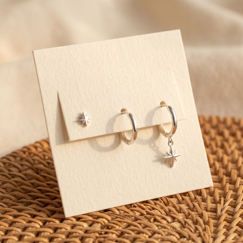 May include: A set of silver-toned jewelry on a cream-colored card, featuring one small star-shaped stud earring, one plain hoop earring, and one hoop earring with a dangling star-shaped charm, all with a polished metallic finish.