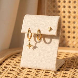 May include: A set of gold-toned jewelry consisting of two hoop earrings and one stud earring. One hoop features a dangling eight-pointed star charm, while the other hoop is plain. The stud earring is shaped like a small eight-pointed star.