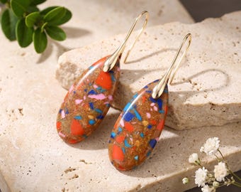 Puede incluir: Un par de pendientes colgantes en forma de lágrima con una base marrón y motas multicolores incrustadas en tonos rojo, azul y rosa. Los pendientes están sujetos a ganchos de metal dorado que salen de la parte superior de cada colgante.