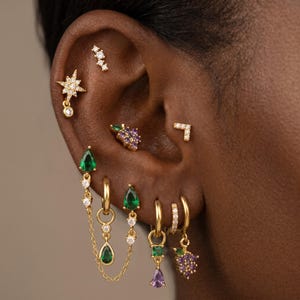 Può includere: Una collezione di orecchini color oro con vari design, tra cui una stella con una pietra pendente, un gruppo di pietre viola, una forma a chevron e diversi cerchi con pietre verdi e viola a forma di goccia collegate da catenelle.