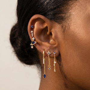 May include: A collection of gold-tone earrings featuring celestial motifs including stars, a planet with rings, and small blue and clear stones. The set includes a vertical climber earring, a hoop with a baguette drop, and two chain-link dangle earrings.