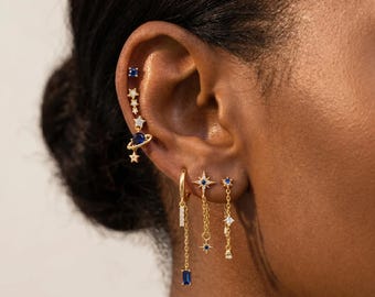 May include: A collection of gold-tone earrings featuring celestial motifs including stars, a planet with rings, and small blue and clear stones. The set includes a vertical climber earring, a hoop with a baguette drop, and two chain-link dangle earrings.