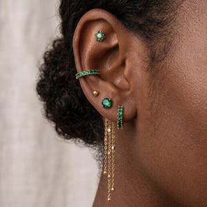 以下が含まれることがあります： 緑色の石があしらわれたゴールドトーンのピアスコレクション。花の形をしたスタッド、小さなフープ、丸いスタッド、シンプルなボールスタッド、そして小さなゴールドのビーズが付いた3本の繊細なチェーンが揺れるフープピアスが含まれています。