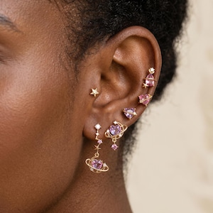May include: A collection of gold-tone earrings featuring pink heart-shaped and teardrop-shaped stones. The designs include celestial motifs like stars and rings resembling planets, with small clear accent stones set along the metal structures.