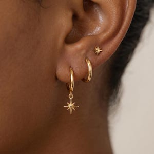 Puede incluir: Un conjunto de tres pendientes dorados que consta de un pequeño perno en forma de estrella, un aro liso y un aro con un dije de estrella colgante. Cada pieza tiene un acabado pulido y un diseño de estrella con múltiples rayos.