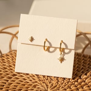 May include: A set of three gold-toned earrings displayed on a cream-colored card. The set includes one small stud earring shaped like a star, one plain gold hoop earring, and one gold hoop earring featuring a dangling star-shaped charm.