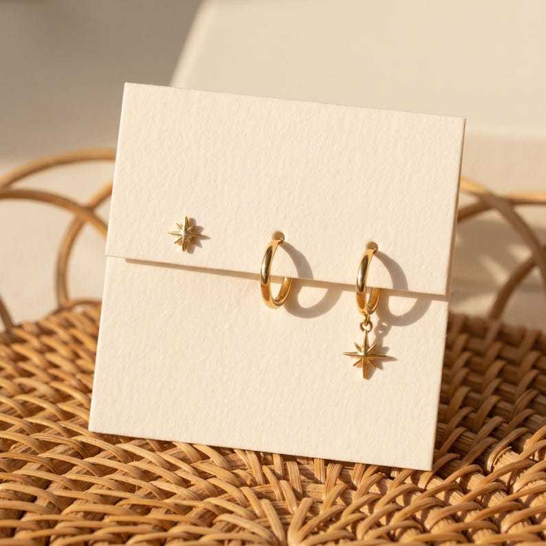 May include: A set of three gold-toned earrings displayed on a cream-colored card. The set includes one small stud earring shaped like a star, one plain gold hoop earring, and one gold hoop earring featuring a dangling star-shaped charm.