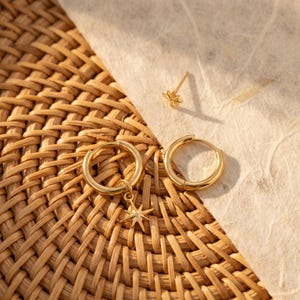 May include: A set of gold-toned jewelry consisting of two hoop earrings and one stud earring. One hoop features a dangling eight-pointed star charm, while the other hoop is plain. The stud earring is shaped like a small eight-pointed star.