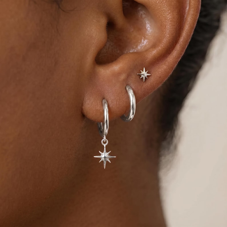 May include: A set of three silver-toned earrings consisting of one small stud earring shaped like a four-pointed star, one plain silver hoop earring, and one silver hoop earring featuring a dangling four-pointed star charm pendant.