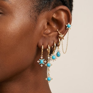 May include: A set of gold-toned earrings featuring turquoise stones and clear crystals. The collection includes a star-shaped ear cuff with a dangling chain, a hoop with a teardrop charm, a sunburst charm, and a cross-shaped charm with a central stone.