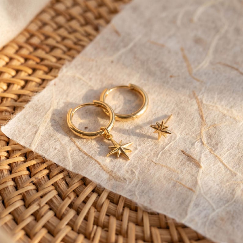 May include: A pair of gold hoop earrings featuring dangling starburst charms and a single matching starburst stud earring. The jewelry pieces are polished with a bright metallic finish, showcasing a classic celestial design with pointed rays.