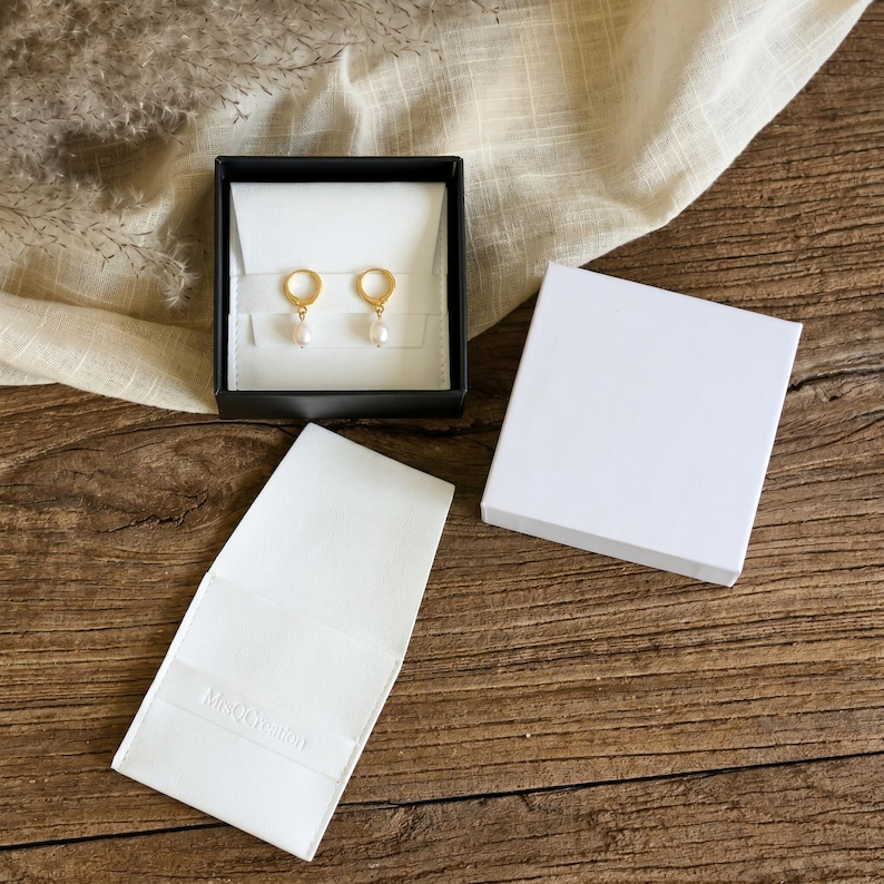 May include: A pair of gold hoop earrings with small white pearl drops displayed inside an open black jewelry box with a white insert. A separate white jewelry box lid and a white paper sleeve with the embossed text MsQCreation are included.