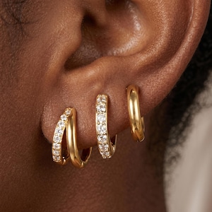May include: Three gold hoop earrings worn in a row on an earlobe. The first earring is a double hoop with one band featuring clear pavé stones. The second is a single hoop with clear pavé stones. The third is a plain, smooth gold hoop earring.