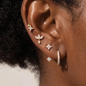 May include: A set of five gold-toned earrings worn in a single ear. The collection includes a star-shaped stud, a marquise-cut floral stud, a four-petaled flower stud, a small round solitaire stud, and a thin hoop earring encrusted with clear stones.