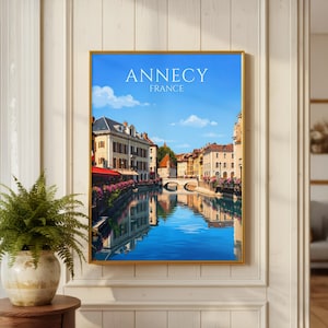 May include: A framed wall art print featuring a scenic view of Annecy, France, with buildings along a canal reflecting in the blue water under a clear sky. The text ANNECY FRANCE is printed in white at the top of the rectangular illustration.