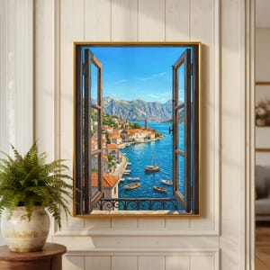 Op de afbeelding: Een ingelijste kunstdruk met een open raamuitzicht op een kuststad met daken van terracotta, een blauwe baai, diverse kleine boten en verre bergen onder een heldere lucht, weergegeven op een witte wand met een varenplant in een pot.