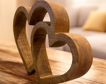 Op de afbeelding: Een houten sculptuur van twee in elkaar verweven harten. De harten zijn gesneden uit lichtbruin hout en hebben een glad, gebogen ontwerp. De sculptuur staat op een houten oppervlak, met een vage achtergrond.