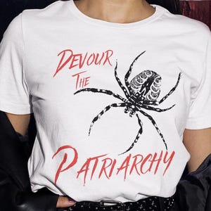 Puede incluir: Camiseta blanca con un diseño de araña en blanco y negro y el texto rojo "Devour the Patriarchy". La araña tiene patas detalladas y un cuerpo estampado. Prenda de vestir casual.