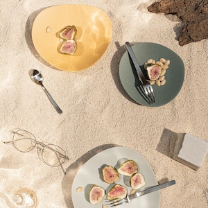 May include: Overhead shot of three irregularly shaped plates in yellow, green, and light blue, each holding sliced figs. A silver spoon, a pair of glasses, and a fork are also present on the sandy surface. The scene evokes a sense of a relaxed meal.