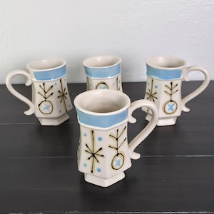 May include: A set of four off-white ceramic mugs with a hexagonal base and a light blue band around the rim. Each mug features a hand-painted pattern of black starburst lines, small light blue dots, and circular motifs with blue crosses.