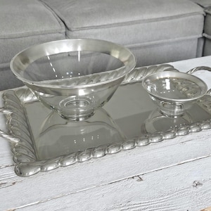 May include: A rectangular silver-toned serving tray with a rope-style border and two handles, featuring a large clear glass bowl with a silver rim and a smaller matching clear glass bowl with a silver rim positioned on the tray surface.