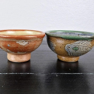 May include: Two ceramic bowls with a footed base. The left bowl is orange-brown with a light interior and a fish illustration. The right bowl is tan with a green rim and a detailed fish graphic. Both bowls feature a textured, speckled finish.