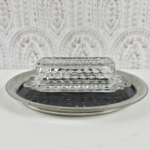 May include: A clear glass butter dish with a diamond-patterned lid and base, resting on a round silver-toned metal tray with a beaded edge. The butter dish features small handles on each side and a textured surface that catches the light.