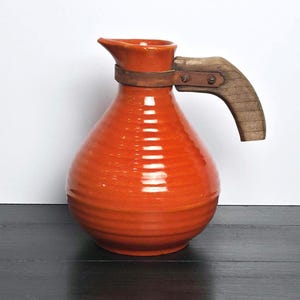 May include: A bright orange ceramic pitcher with a bulbous, ribbed body and a narrow neck. The pitcher features a curved, textured wooden handle attached to the neck by a flat metal band secured with two visible screws on the side.