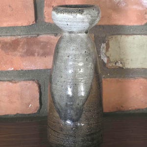 May include: A handmade ceramic vase with a unique shape and a speckled, glazed finish. The vase features a light gray top and a darker gray body, with a brown base. The vase is approximately 7 inches tall.