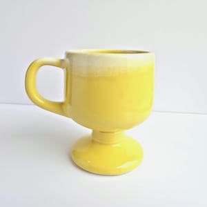 May include: A yellow ceramic mug with a rounded base and a handle. The mug has a white rim and a pedestal base. The mug is a bright yellow color.