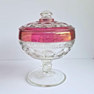 May include: A clear glass candy dish with a lid. The rim of the dish and lid are a vibrant pink color. The dish has a pedestal base and is decorated with a repeating pattern of circles.